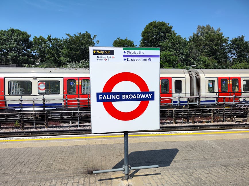 A railway station platform featuring a prominent white sign with a red circle and blue text reading 'Ealing Broadway,' indicating the station name. The sign also displays directions to the 'Way out,'